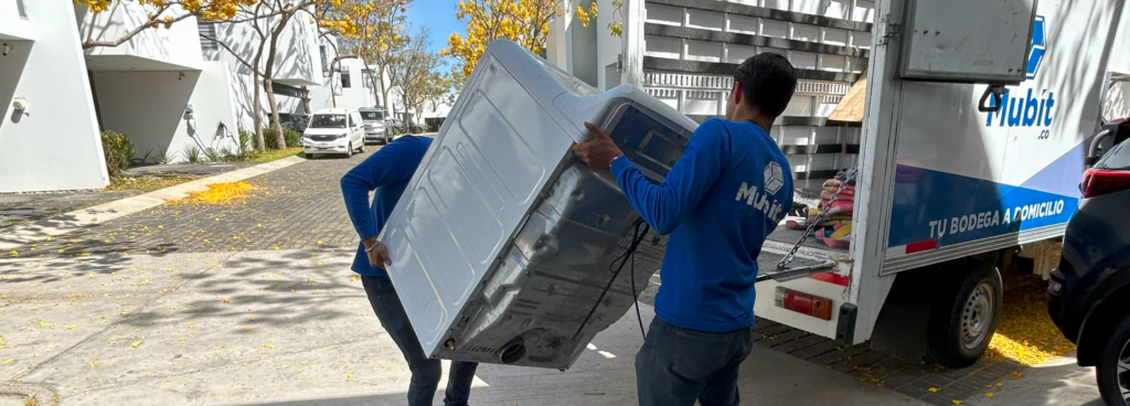 dos hombres vestidos de azul cargando una lavadora para subirla a la parte posterior de una camioneta. Bodega Guadalajara