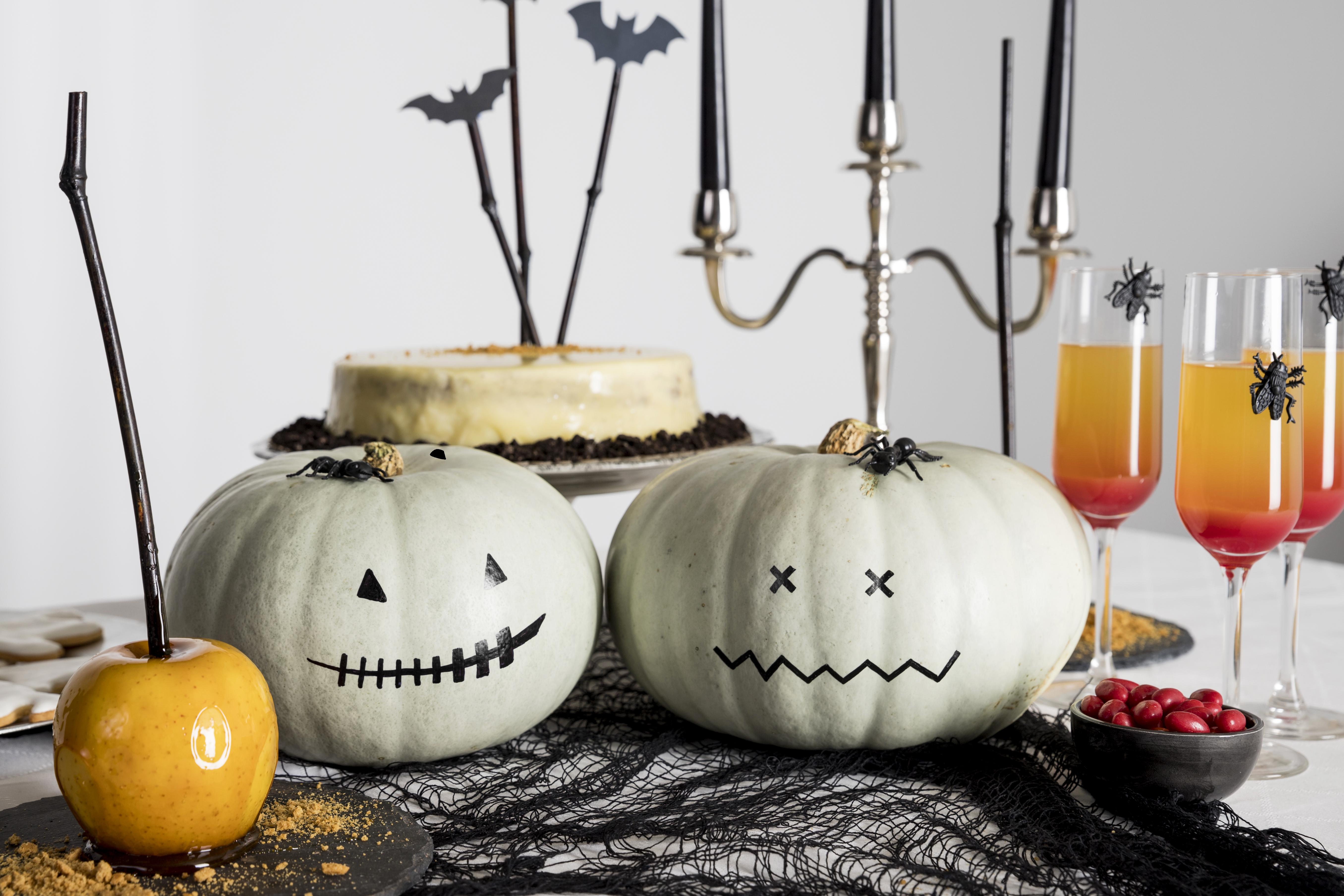 Dos calabazas blancas con caras de Halloween pintadas con plumón, una manzana con caramelo, bebidas en copas, un pastel y velas negras Bodega Guadalajara